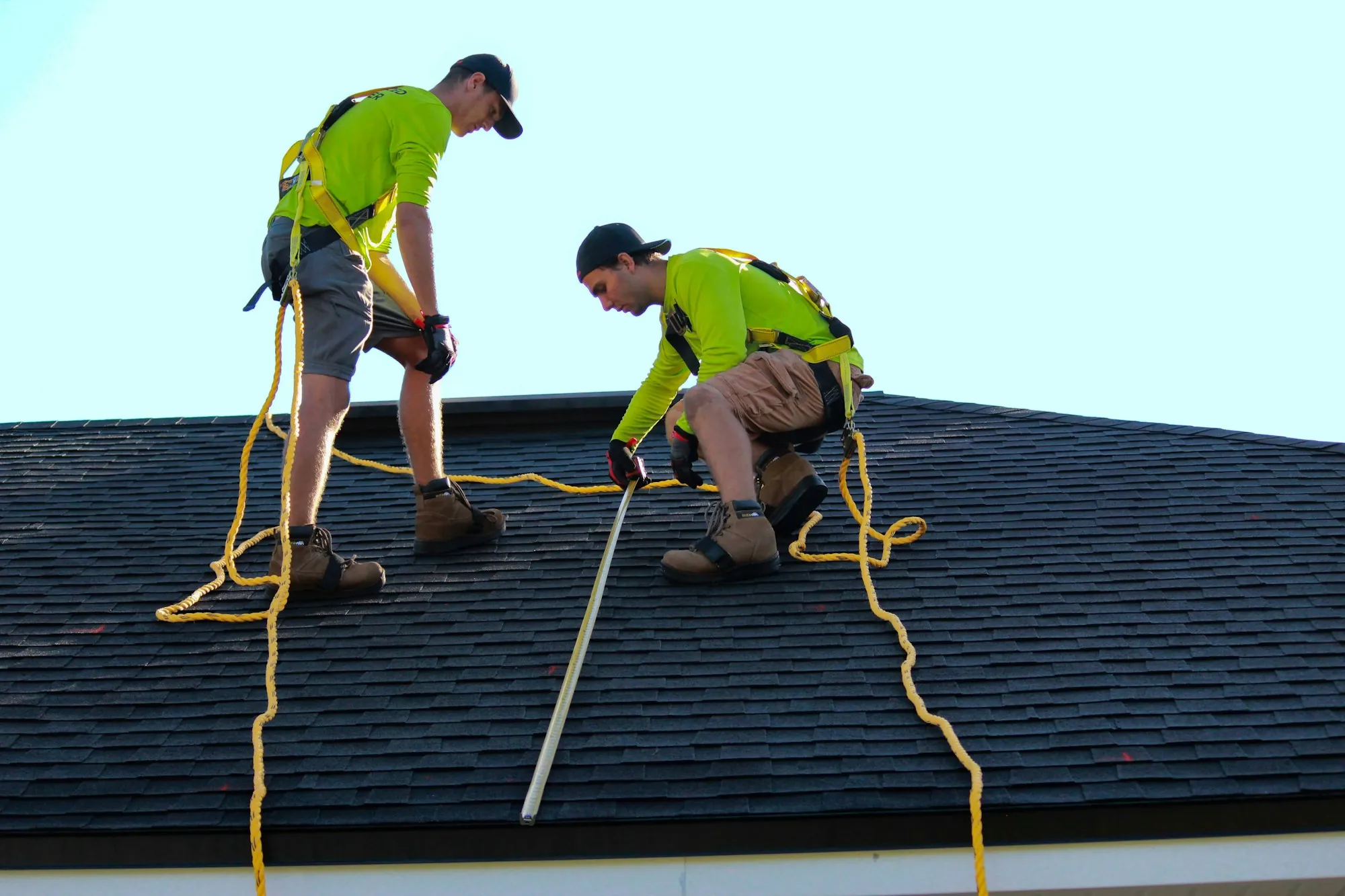 Two roofers in safety harnesses and hi-vis shirts measuring on a dark shingle roof