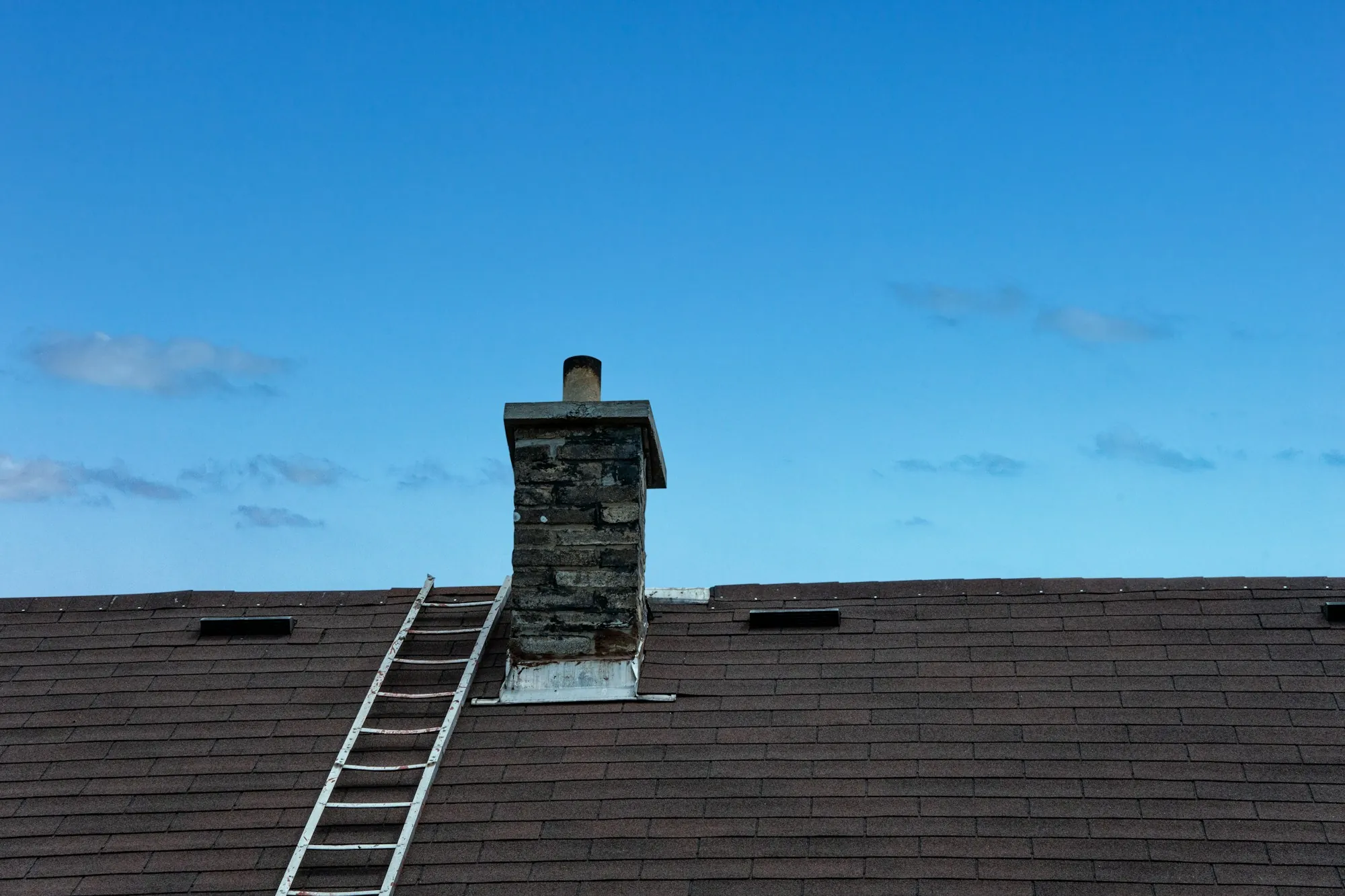 Ladder resting on a dark shingle roof beside a stone chimney
